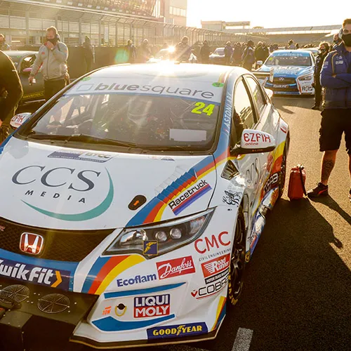 Racing car number 24 on the track with crew members standing by it, surrounded by other vehicles and personnel in the background.