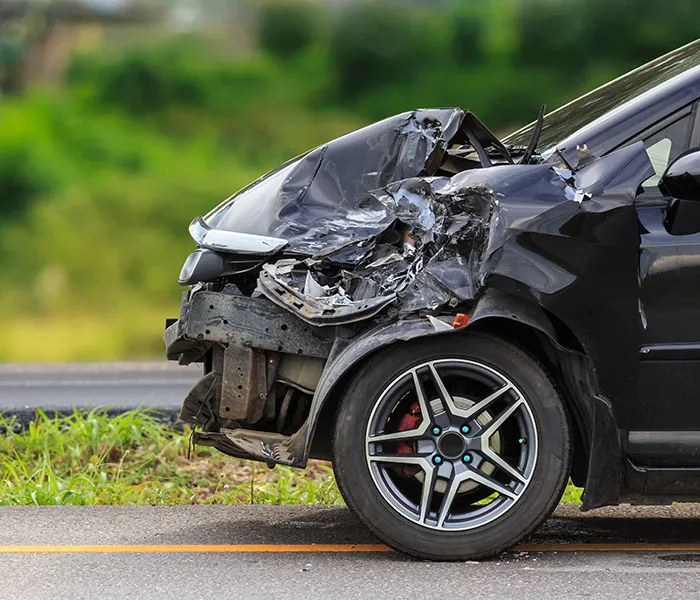 Front-side of a black car heavily damaged from a collision, with a crumpled hood and exposed internals, parked on the roadside.