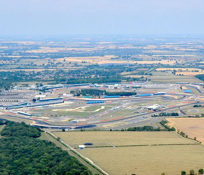 Aerial view of a large motor racing circuit with surrounding fields and countryside under a clear sky.