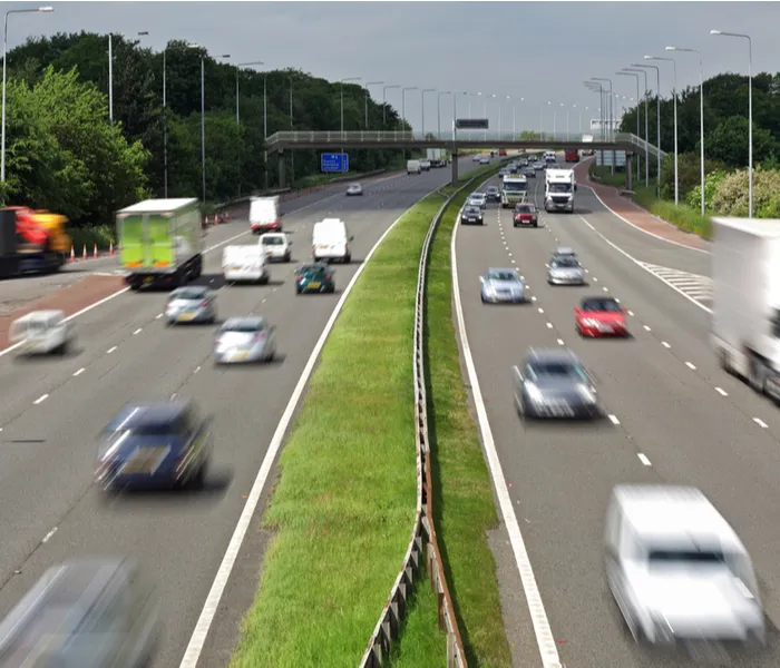 A busy multi-lane highway with various vehicles in motion, a central grass divider, and overhead signs in the background.