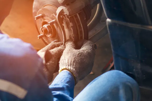 A person wearing gloves and blue overalls replaces a brake pad on a vehicle's wheel assembly in a workshop setting.