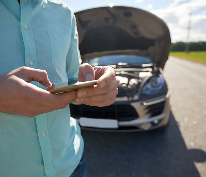 Person in a light blue shirt using a smartphone with a car, hood open, parked on the roadside in the background.