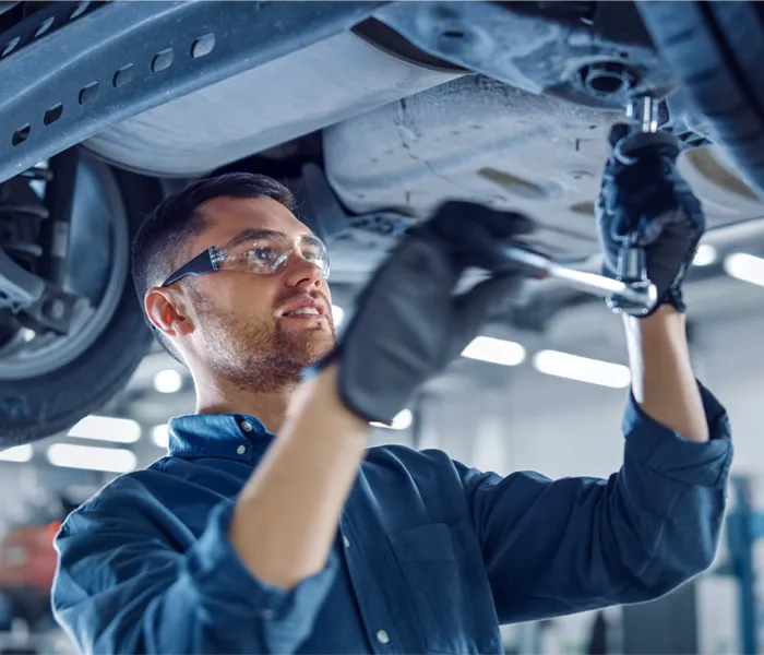 Mechanic wearing safety glasses and gloves uses a wrench to work under a vehicle in a well-lit auto repair shop.