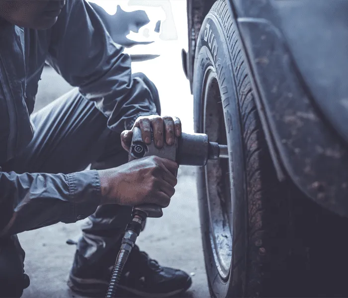 A person in work clothing uses a power tool to tighten bolts on the wheel of a vehicle in a garage.