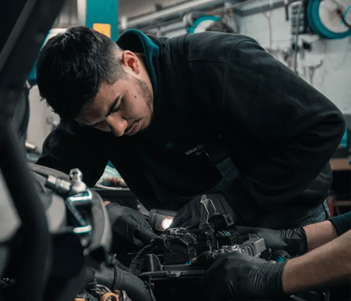 A mechanic in dark clothing and gloves works on a car engine in a garage, focusing intently on the task at hand.