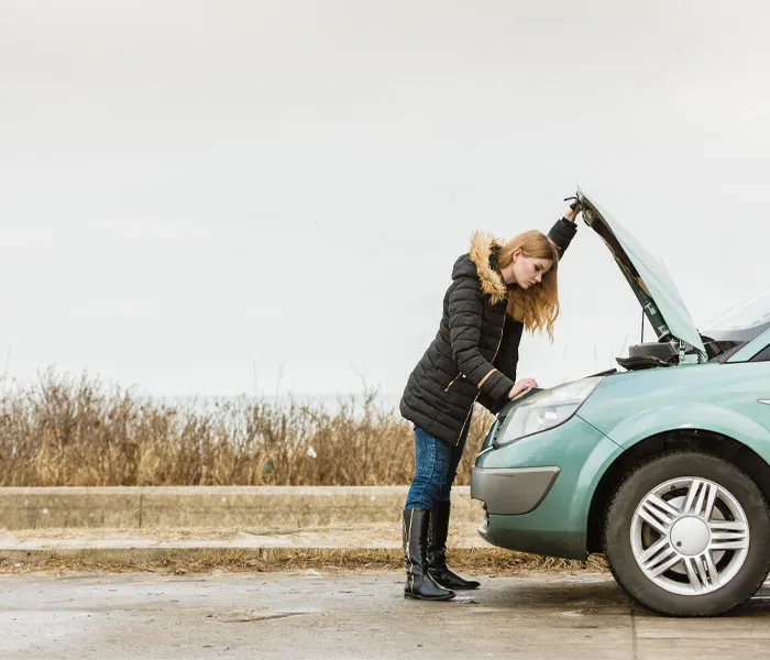 A woman in winter clothing inspects the engine of her green car with the hood open, parked on a roadside.