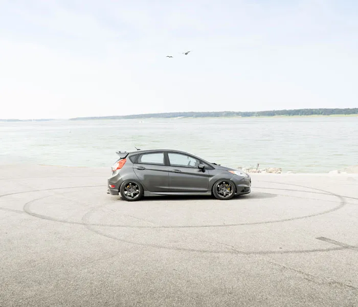 A parked black compact car is located on a stretch of pavement near a large body of water with trees in the background.