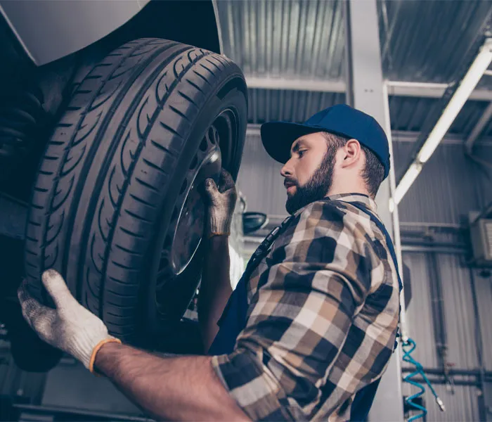 Mechanic wearing a plaid shirt and cap, adjusting a car tire on a raised vehicle in an automotive repair shop.