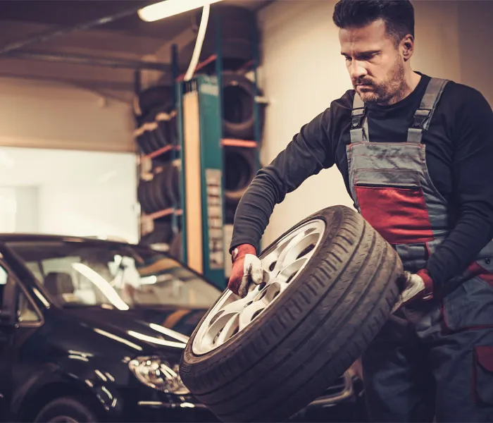 Mechanic in a garage holding a car tire, with a black car in the background and shelves of tires behind him.