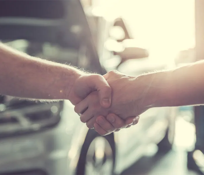 Two people shaking hands with cars in the background, suggesting a deal or agreement in a well-lit environment.